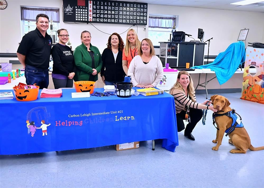  A group of people, including a working dog, take a photo in front of a table.